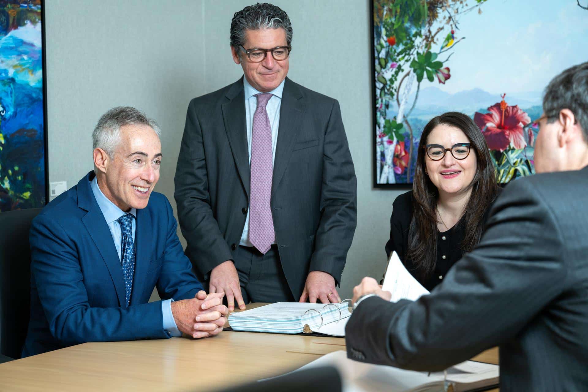 Image of an Elman Freiberg Partners meeting between Jay W. Freiberg, Howard I. Elman, Yelena Rapoport, and the back of an unidentifiable counsel member (left to right). Smiling and sitting around conference room table looking over documents in binders.