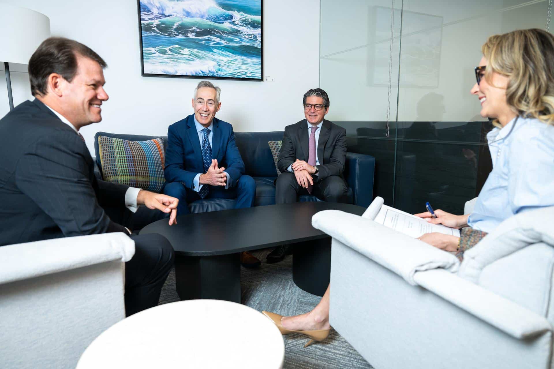 Image of an Elman Freiberg Partners meeting between Benjamin S. Litman, Jay W. Freiberg, Howard I. Elman, and Nora Feher (left to right). Smiling and sitting on chairs and couch around coffee table.